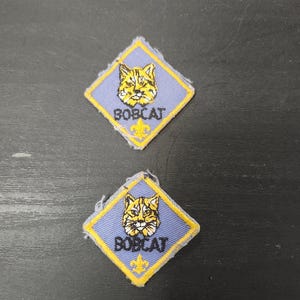 May include: Two blue and yellow Bobcat patches. Each patch features a yellow bobcat head above the word "BOBCAT" in white text. A yellow fleur-de-lis is below the text. The patches are diamond-shaped.