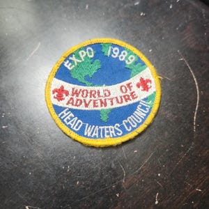 May include: A circular patch with a yellow border, featuring the text "EXPO 1989" and "WORLD OF ADVENTURE HEAD WATERS COUNCIL." The design includes a blue background with a green map of the world and red accents.