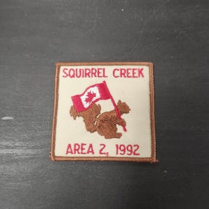 May include: Square patch with the text "SQUIRREL CREEK" above a brown squirrel holding a Canadian flag. The text "AREA 2, 1992" is below the squirrel. The patch has a tan background and a brown border.