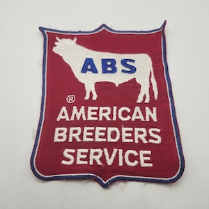 May include: A shield-shaped embroidered patch with a maroon background and a blue border. It features a white silhouette of a bull with the blue text ABS centered on its body. Below the bull, the white text reads AMERICAN BREEDERS SERVICE.