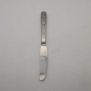 May include: A silver butter knife with a decorative handle featuring a detailed embossed design. The blade is smooth and curved, and the handle has a small, rectangular detail near the blade.