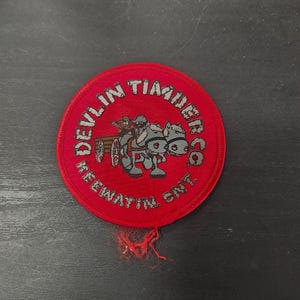 May include: A round, red patch with the text "Devlin Timber Co. Keewatin, Ont." in white lettering. The patch features a cartoon illustration of a person driving a horse-drawn cart. The patch has a frayed edge.