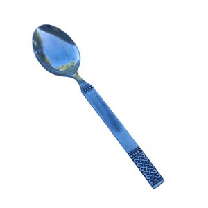 May include: A polished silver-tone metal spoon featuring a smooth, oval-shaped bowl and a flat, straight handle. The handle is decorated with two distinct bands of dark, etched geometric patterns and a series of small dots near the neck.