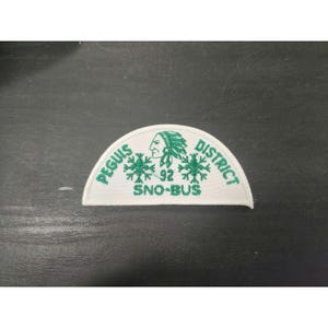 May include: A white, semi-circular patch with green embroidered text and graphics. The text reads "PEGUIS DISTRICT" and "SNO-BUS 92". A profile of a Native American person is in the center, with snowflake-like designs on either side.