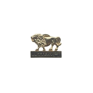 Puede incluir: Un pin de león dorado con la palabra "LUTTER" y la fracción "1/3" en una base rectangular. El león está en una pose rugiente, con melena y cuerpo detallados. El pin está sobre un fondo blanco.