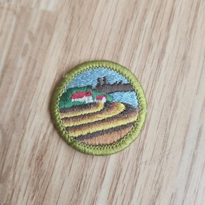 May include: Embroidered patch featuring a landscape scene with a blue sky, water, and green and brown fields. The patch has a green border and depicts small houses with red roofs. The patch is round.