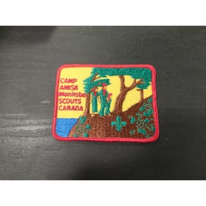 May include: Embroidered patch with a yellow background featuring a green tree and two figures. The patch has the text "CAMP AMISK Manitoba SCOUTS CANADA" in white. The patch is bordered in red.
