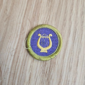 May include: A round embroidered patch featuring a gold lyre symbol on a purple background. The lyre is surrounded by a gold border. The patch is likely for a uniform or craft project.