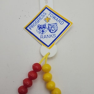 May include: A vintage Boy Scout Progress Toward Ranks badge. The white plastic badge has a blue and yellow diamond-shaped emblem with text and animal illustrations. Red and yellow beads hang from the badge.