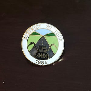 May include: A round pin with the text "DISTRICT 30 TOUR" in white lettering. The pin features a road scene with a motorcycle and the text "AMA 1985". The pin has a white border and a dark background.