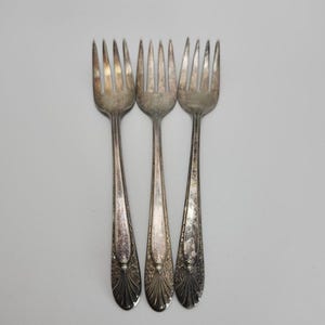 May include: Three antique silver forks with ornate handles. The forks are arranged vertically against a white background. The tines are slightly tarnished, and the handles feature a detailed floral design.