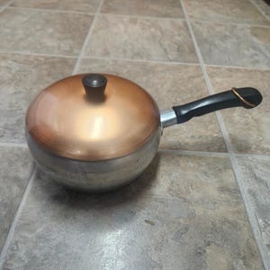 May include: A round cooking pot with a metallic silver base and a polished copper-colored lid. The lid features a small, dark brown knob handle. A black handle extends from the side of the pot, equipped with a small metal loop for hanging.