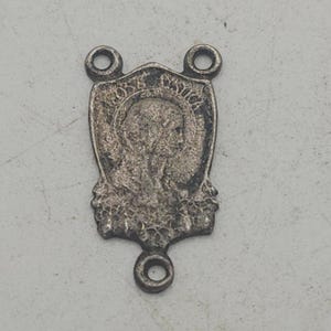 May include: A silver-toned pendant with a shield shape, featuring a central image of a person and the inscription "NO. DOM. JUSTICA". The pendant has two loops at the top and one at the bottom.