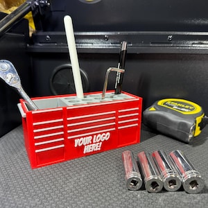 May include: A red toolbox-style desk tidy with white stripes and the text "YOUR LOGO HERE!". It contains tools such as a ratchet, Allen key, and pens. A yellow and black tape measure and four silver sockets are also visible.