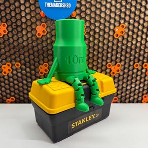 May include: A green 3D printed figurine of a person sitting on a yellow and black Stanley Jr. toolbox. The figurine has a 10mm diameter and is wearing a toolbox as a hat.