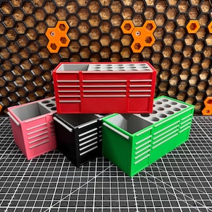 May include: Four small, colourful tool chest organisers in red, pink, black, and green. Each chest has a rectangular shape with white horizontal stripes and a top section with multiple circular holes. The background features a honeycomb pattern.