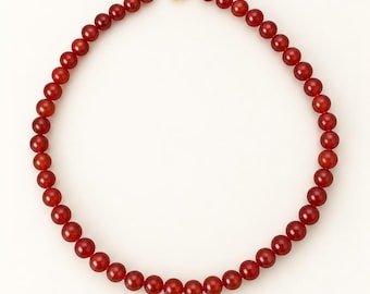 May include: A necklace composed of uniform, smooth, deep red spherical beads arranged in a circular pattern. The strand is secured at the top with a polished gold-toned metal spring ring clasp, providing a secure closure for the beaded jewelry piece.