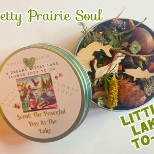 May include: A round tin container with a green lid and a floral design. The tin is labeled "Pretty Prairie Soul" and "A Dreamy Little Lake Flower Soup To-Go". The scent is "The Peaceful Day At The Lake".
