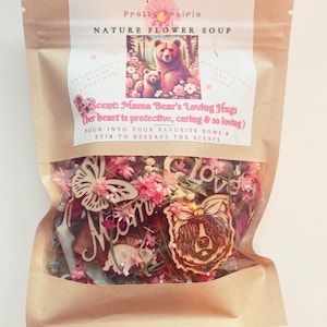 Könnte beinhalten: Ein brauner Papierbeutel mit einem Etikett, auf dem "Pretty Prairie Nature Flower Soup" steht. Das Etikett zeigt eine Comic-Mamabärin und ein Junges mit dem Text "Scent: Mama Bear's Loving Hugs (Her heart is protective, caring & so loving)". Der Beutel enthält getrocknete Blumen, Holzausschnitte und einen Holzausschnitt einer Mamabärin mit dem Text "Mama Bear".