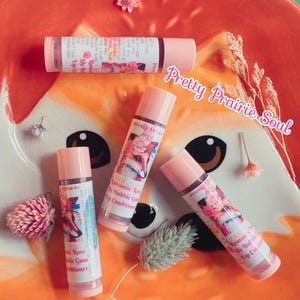 Puede incluir: Varios acondicionadores labiales rosas se muestran en un plato naranja. Los bálsamos labiales tienen etiquetas con una ilustración de zorro y el texto "Pretty Prairie Soul". El texto "Romantic Rose Shy Bubble Gum Lip Conditioner" también es visible.