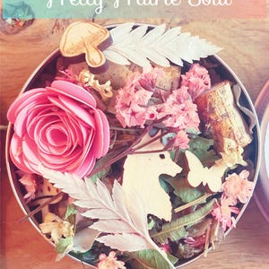 Puede incluir: Una lata redonda de metal llena de flores secas, hojas y formas de madera. La lata contiene una rosa rosa, un helecho blanco, un hongo de madera, una mariposa de madera y una forma de unicornio de madera. El texto "Pretty Prairie Soul" está escrito en rosa y azul sobre un fondo azul claro en la parte superior de la imagen.