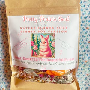 Puede incluir: Una bolsa de papel marrón con una etiqueta blanca que dice "Pretty Prairie Soul" y "Nature Flower Soup Simmer Pot Version". La etiqueta también incluye una ilustración vintage de un conejito rosa en un entorno de bosque. El aroma se describe como "Easter in the Beautiful Forest" e incluye una lista de ingredientes: zanahorias, capullos de rosa, pomelos, pino, coco, salvia, etc. La bolsa contiene una mezcla de hierbas y flores secas.