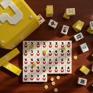 Puede incluir: Una caja amarilla con un signo de interrogaci&oacute;n blanco y una colecci&oacute;n de fichas de juego. Las fichas son blancas con n&uacute;meros negros, caracteres chinos y s&iacute;mbolos 'W' rojos. Algunas fichas son amarillas con signos de interrogaci&oacute;n.