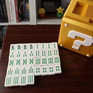 Puede incluir: Un conjunto de fichas de Mahjong blancas con marcas verdes se organiza sobre una superficie marr&oacute;n oscuro. Un gran bloque dorado con un signo de interrogaci&oacute;n est&aacute; cerca. El bloque tiene un signo de interrogaci&oacute;n blanco en la parte delantera.