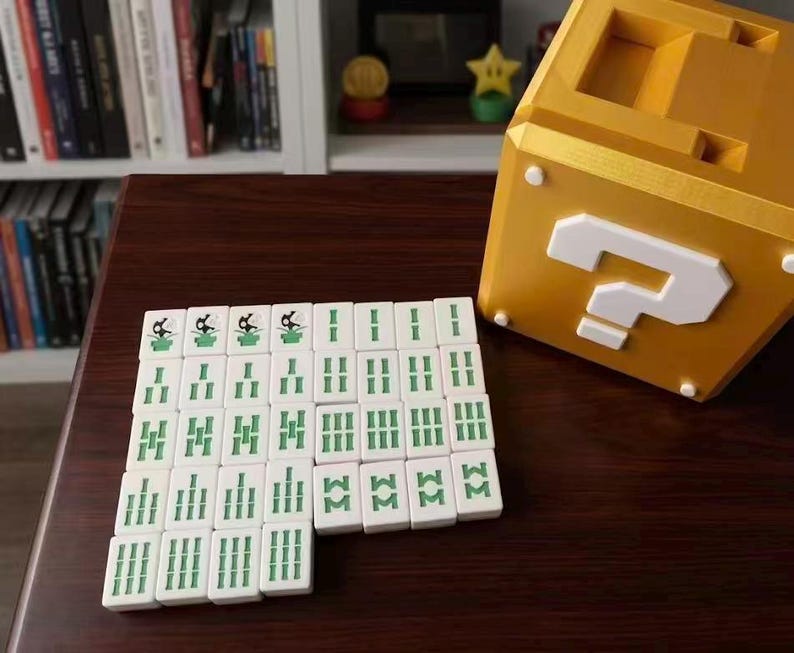 Puede incluir: Un conjunto de fichas de Mahjong blancas con marcas verdes se organiza sobre una superficie marr&oacute;n oscuro. Un gran bloque dorado con un signo de interrogaci&oacute;n est&aacute; cerca. El bloque tiene un signo de interrogaci&oacute;n blanco en la parte delantera.