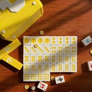 Puede incluir: Un juego de mesa con fichas blancas y s&iacute;mbolos amarillos, as&iacute; como una caja amarilla. Tambi&eacute;n hay monedas y dados amarillos, todo dispuesto sobre una superficie de madera.