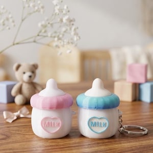 May include: Two white baby bottle-shaped keychains, one with a pink top and the other with a blue top. Each keychain has a heart with the word "MILK" on it. A silver keyring is attached to the blue keychain. The background includes a teddy bear and wooden blocks.
