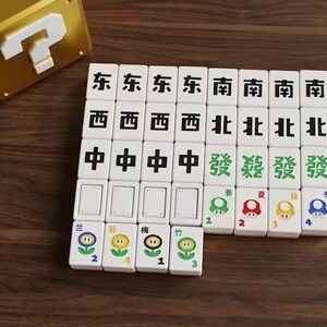 Puede incluir: Un juego de fichas de Mahjong blancas con caracteres chinos negros e ilustraciones coloridas con tem&aacute;tica de Mario. Una caja dorada con un signo de interrogaci&oacute;n blanco est&aacute; en el fondo. Las fichas est&aacute;n dispuestas sobre una superficie de madera marr&oacute;n.