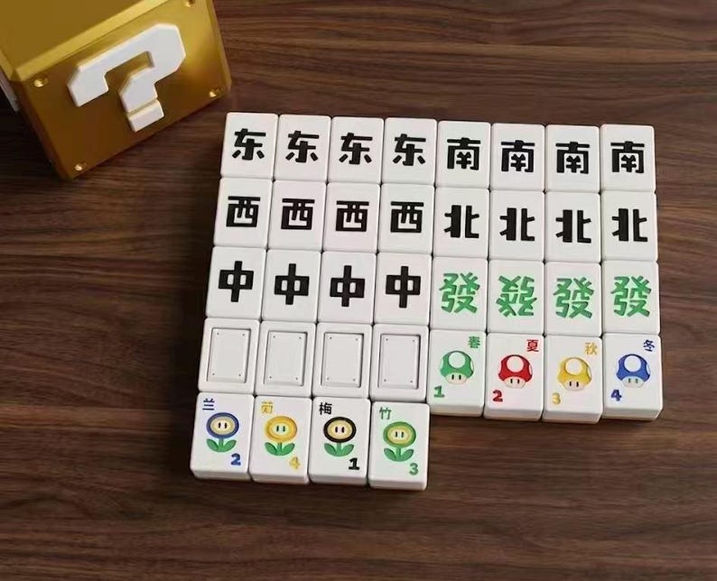 Puede incluir: Un juego de fichas de Mahjong blancas con caracteres chinos negros e ilustraciones coloridas con tem&aacute;tica de Mario. Una caja dorada con un signo de interrogaci&oacute;n blanco est&aacute; en el fondo. Las fichas est&aacute;n dispuestas sobre una superficie de madera marr&oacute;n.