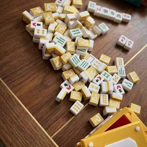 Puede incluir: Un conjunto de fichas de Mahjong de color marfil y dorado dispersas sobre una superficie de madera. Las fichas muestran varios s&iacute;mbolos y n&uacute;meros en rojo, verde y negro. Una caja amarilla con un gran n&uacute;mero 3 blanco est&aacute; en primer plano, sugiriendo un juego.