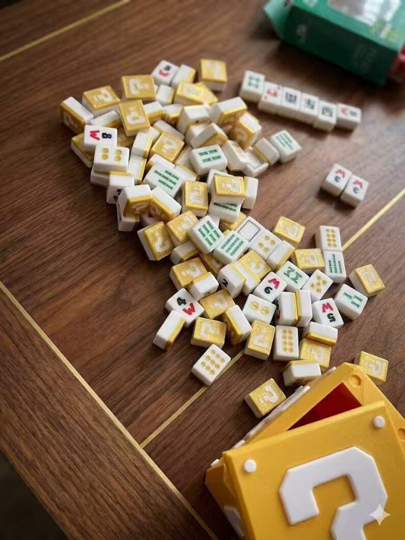 Puede incluir: Un conjunto de fichas de Mahjong de color marfil y dorado dispersas sobre una superficie de madera. Las fichas muestran varios s&iacute;mbolos y n&uacute;meros en rojo, verde y negro. Una caja amarilla con un gran n&uacute;mero 3 blanco est&aacute; en primer plano, sugiriendo un juego.