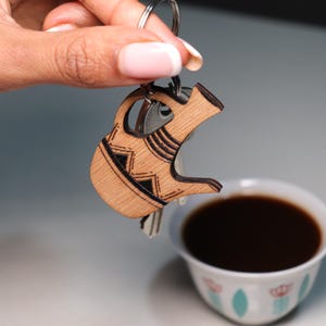May include: A light brown wooden keychain shaped like a traditional coffee pot, featuring dark engraved geometric patterns on its surface. The keychain is attached to a metal ring that holds several silver-colored metal keys.