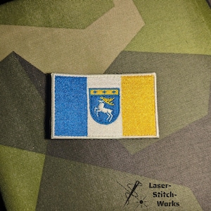 Könnte beinhalten: Gestickter Aufnäher mit Flaggenmotiv. Er hat blaue, weiße und gelbe vertikale Streifen. Die Mitte hat ein blaues Wappen mit einem weißen Hirsch und goldenen Details. Der Aufnäher ist rechteckig und hat einen genähten Rand.