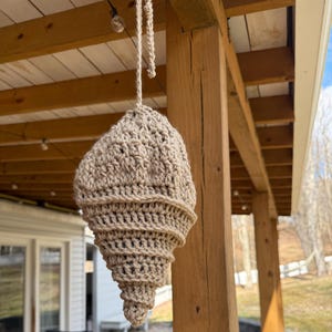 May include: A beige crocheted seashell hanging from a wooden porch ceiling. The seashell is made of yarn and has a spiral shape.