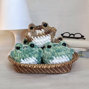 May include: Three crocheted frog plushies in a woven basket. Two frogs are green and white, and one is brown and white. The frogs have black eyes and are made of a soft, textured yarn. The basket is brown.
