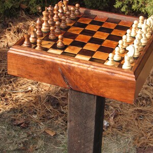 May include: A wooden chess set with a dark brown frame and a checkered playing surface. The chess pieces are light and dark brown. The set is mounted on a wooden post, with the board partially set up for a game.
