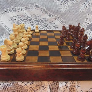 May include: A wooden chess set with light and dark pieces arranged on a checkered board. The chess pieces are in a classic design, with a dark brown wooden frame. The board is set up, ready for a game.