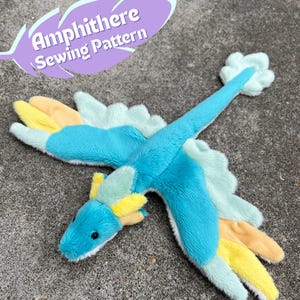 May include: A plush, colorful Amphithere dragon toy with a turquoise body, yellow and orange wings, and a light blue tail. The text "Amphithere Sewing Pattern" is displayed in a purple feather shape.
