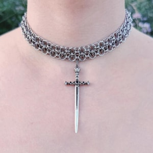 Puede incluir: Un collar gargantilla de cota de malla plateada con un colgante de espada. La gargantilla está hecha de múltiples filas de pequeños anillos metálicos entrelazados. El colgante de espada tiene una empuñadura detallada y una hoja larga y puntiaguda.