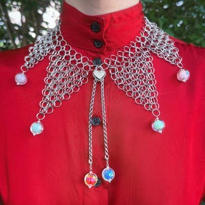 Puede incluir: Un collar de cota de malla plateada con orbes iridiscentes colgantes. El collar tiene un colgante en forma de coraz&oacute;n y se usa sobre una camisa roja con cuello. Los orbes son en tonos rosa, azul y verde.
