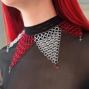 May include: A red and silver chainmail choker necklace with star and crescent moon charms. The necklace is worn by a person with red hair.