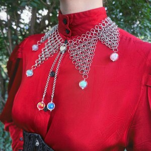 Puede incluir: Un collar de cota de malla plateada con orbes iridiscentes colgantes y un dije en forma de coraz&oacute;n. El collar se usa sobre una blusa roja y un cors&eacute; negro. Una pieza de joyer&iacute;a &uacute;nica.