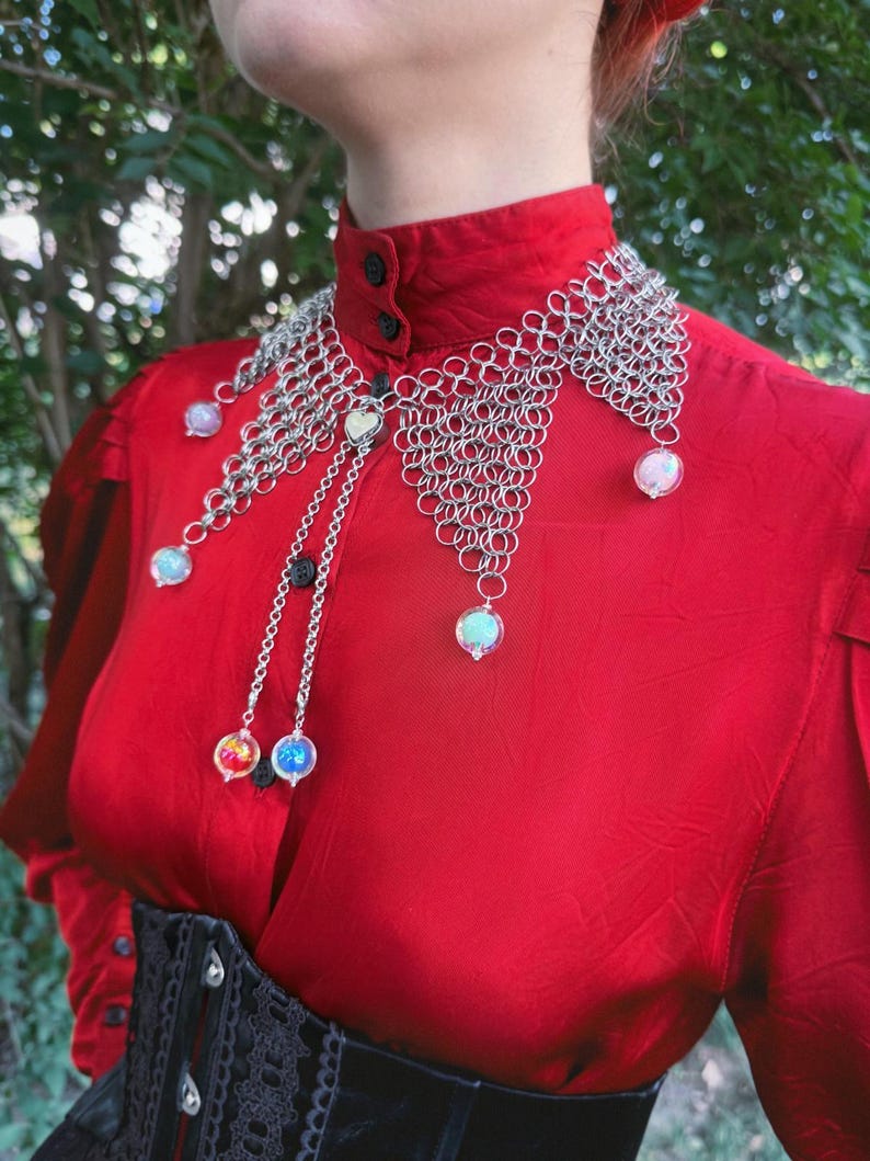 Puede incluir: Un collar de cota de malla plateada con orbes iridiscentes colgantes y un dije en forma de coraz&oacute;n. El collar se usa sobre una blusa roja y un cors&eacute; negro. Una pieza de joyer&iacute;a &uacute;nica.