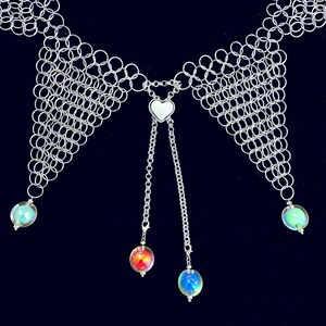 Puede incluir: Un collar de cota de malla plateada con un colgante de coraz&oacute;n y cadenas colgantes. El collar presenta orbes iridiscentes en los extremos de las cadenas y en los lados. El fondo es azul oscuro.