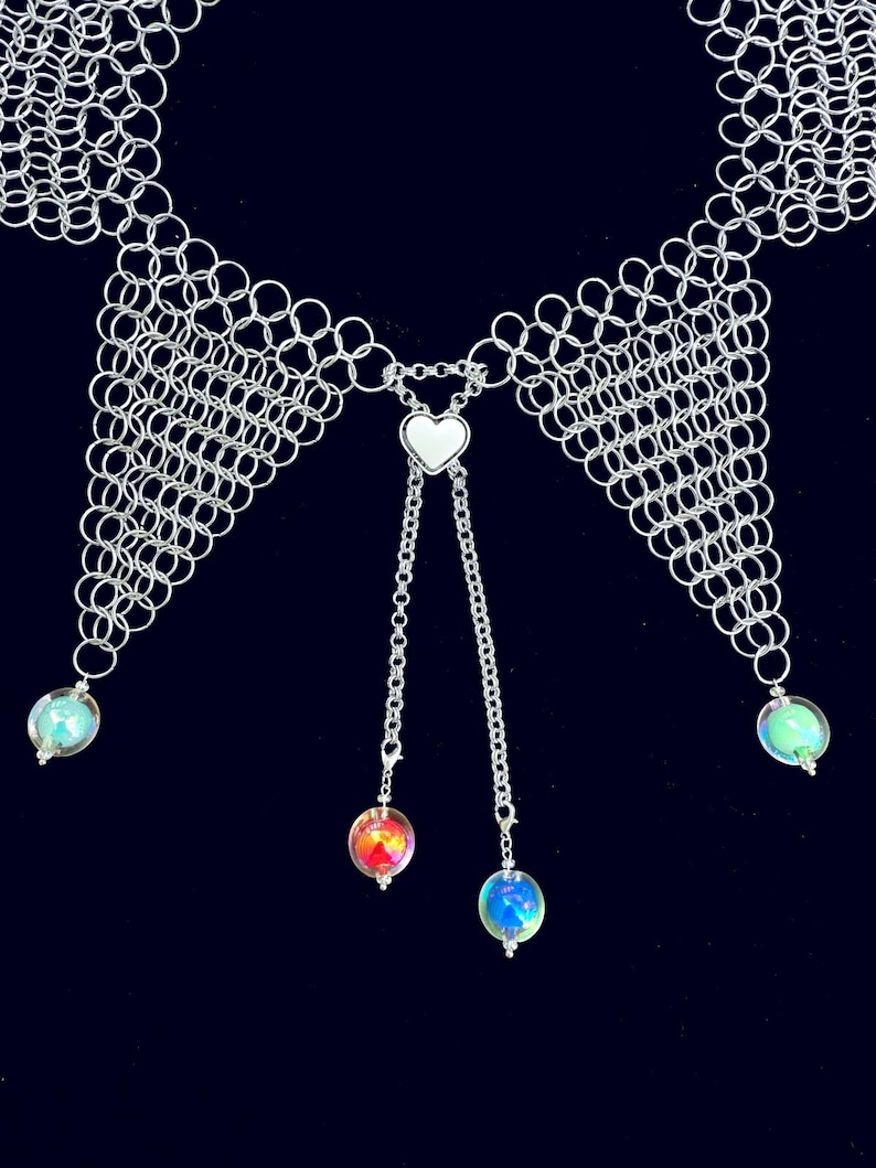 Puede incluir: Un collar de cota de malla plateada con un colgante de coraz&oacute;n y cadenas colgantes. El collar presenta orbes iridiscentes en los extremos de las cadenas y en los lados. El fondo es azul oscuro.
