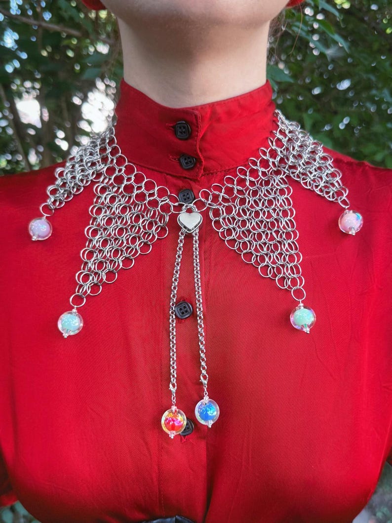 Puede incluir: Un collar de cota de malla plateada con orbes iridiscentes colgantes. El collar tiene un colgante en forma de coraz&oacute;n y se usa sobre una camisa roja con cuello. Los orbes son en tonos rosa, azul y verde.